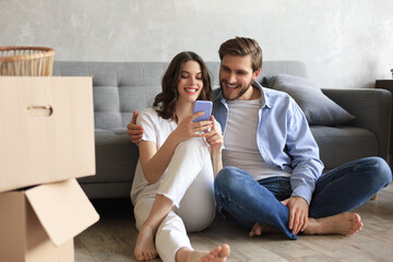 Happy couple sitting and planning new home decoration at home with a smartphone at moving day,...