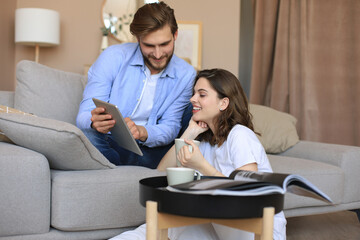 Happy young couple sitting, relaxing on floor in living room watching media content online, doing online shopping in a tablet.