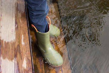 Child in rubber boots stands on wooden flooded bridge and puts his legs in water of river.
