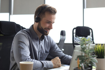 Obraz na płótnie Canvas Smiling male business consultant with headphones sitting at modern office, video call looking at laptop screen. Man customer service support agent helpline talking online chat.