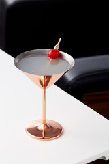 White cloudy cocktail decorated with a cherry on the stick is contained in bronze cocktail glass....