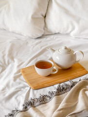 Fototapeta na wymiar Tray of tea on bed. White bedding sheets with blanket and pillow. Breakfast in bed. Warm and cosy scandinavian hygge concept - cup of tea.