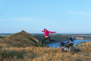 A man in a red sweatshirt with a hood funny jump. Motorcycle driver at the top of the hill overlooking the rocks and the river. Hobbies and leisure. Far from civilization.