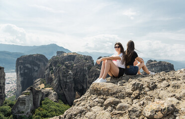 Naklejka na ściany i meble Two sisters sitting at the cliff with breathtaking view at landscape of Meteora, Greece.