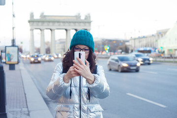 A girl in a down jacket and a hat walks around the city and takes photos on a smartphone,