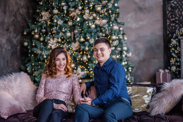 A fashion couple in love sits on a modern sofa in the middle of a dark loft. In the background there is a festive tree decorated with a garland of lights, golden balls, a vintage fireplace with gifts.