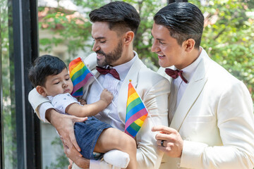 Young gay in white tuxedo is holding adopted son in his hands with LGBT flags at home. Two gays are...