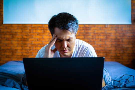 headache, tense young asian man working on laptop computer in bedroom at night