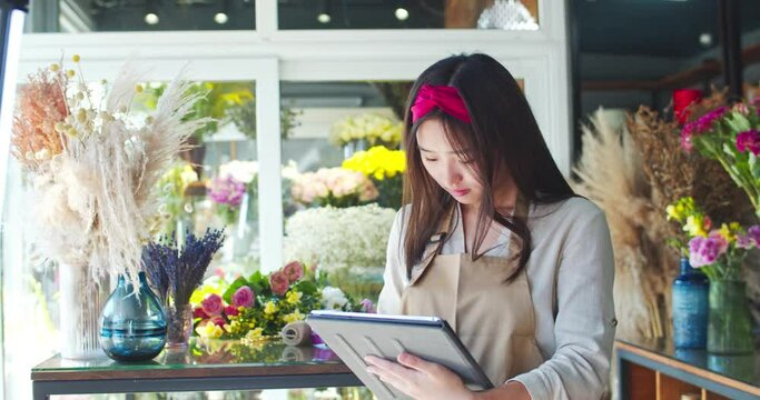 Attractive Asian female store owner wearing apron, working in shop. Happy young woman florist using tablet to take inventory and check availability. Business, nature, entreprenuership concept.
