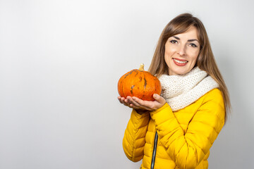 beautiful young girl in a yellow jacket holds a pumpkin on a light background. Halloween concept, celebration