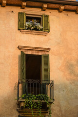 The wall of an old house, on it a balcony with open shutters and flower pots. Above the balcony, a small window with flowers