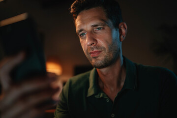 Obraz premium Handsome and confident young businessman in polo t-shirt working at night on smartphone texting message, Professional male entrepreneur typing message or browsing internet looking at cellphone screen