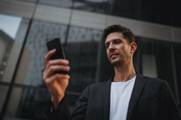 Young successful businessman in casual clothes using smartphone during coffee break near modern office buildings, smiling confident male manager texting email to the customer