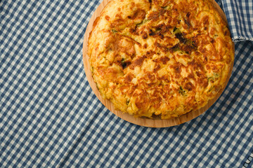 Tortilla de Patatas served on a wood board and a blue checkered tablecloth. Traditional Spanish dish with eggs, potatoes, onion, olive oil and green peppers. 