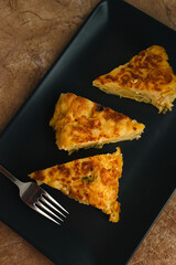 Tortilla de Patatas served on a dark plate. Traditional Spanish dish with eggs, potatoes, onion and green peppers. Rustic black background.