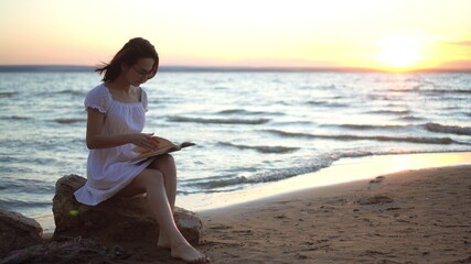 Fototapeta na wymiar A young woman sits on a stone on the beach by the sea with a book in her hands. A girl in a white dress at sunset reads a book.