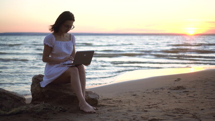 A young woman sits on a stone on the beach by the sea with a laptop in her hands. A girl in a white dress at sunset is chatting in a laptop.