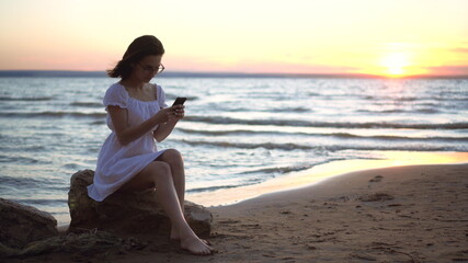 Fototapeta na wymiar A young woman sits on a stone on the beach by the sea with a phone in her hands. A girl in a white dress at sunset.