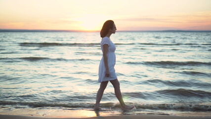 Attractive young woman walks along the sea beach at sunset. A girl in a white dress walks barefoot along the beach.