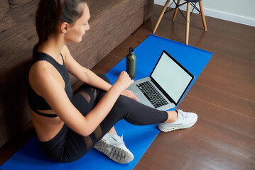 A sideways photo of a sporty girl in black tight pants and top is sitting on the blue yoga mat with a computer and a bottle at home. A mockup of a laptop and fitness woman.