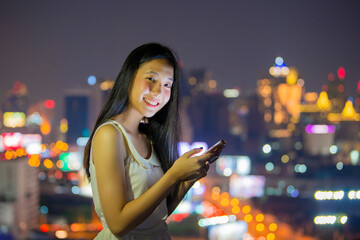 Beautiful girl using smartphone outdoors, Girl chatting with friends at social networks while standing on rooftop balcony with Night blurred bokeh light city office building.