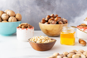 Different nuts (pine nuts, walnuts, hazelnuts and almonds) in beautiful wooden and ceramic bowls stand on a white table on a gray background. There is a small jar of honey on the right