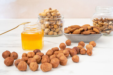 Peeled hazelnuts lie on a white table. Behind it is a small jar of honey, a rectangular gray bowl of peeled almonds, and two glass jars of inshell pistachios and peeled walnuts