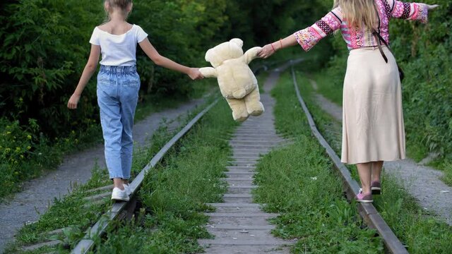 Mom and daughter holding teddy bear walking on train rails at railway tracks. Woman together teenager girl walking on metal railroad rail with teddy bear