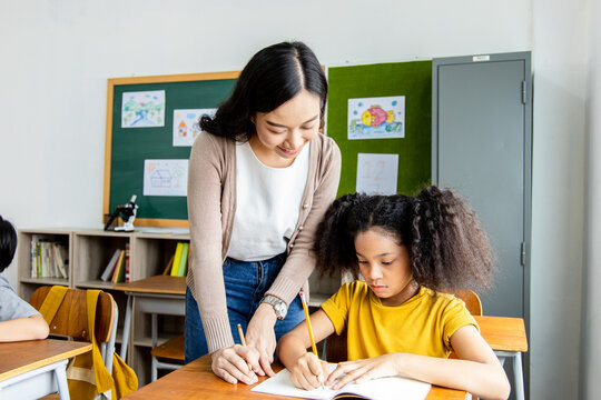 An Asian woman teacher teaches an African American child student a lesson in the classroom. They smile happily during the lessons learned. School diversity student