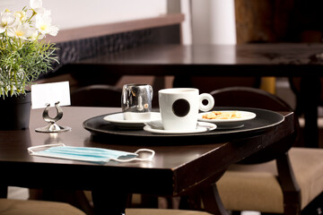 Freshly brewed hot white coffee on the wooden table inside the cafeteria with protection mask, covid-19, cookies, and metal milk jug and free space for decoration.