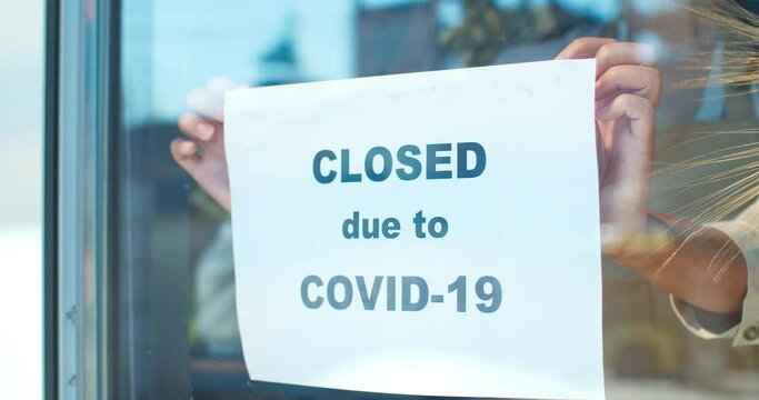 Young female store owner closing shop during epidemic period. Good-looking woman florist in mask attaching paper with text "closed due to COVID-19" to window. Pandemic of coronavirus.