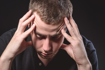 Fototapeta premium Young handsome man in a black shirt sits and holds his head on a black background. Experiencing stress, pain