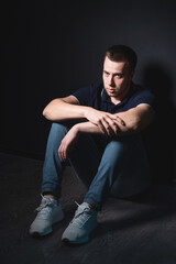 Young attractive man in blue jeans and a polo shirt sits on the floor near the wall in the studio. Youth and attractiveness concept