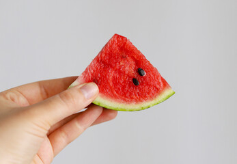 A small slice of red ripe watermelon with black seeds in a woman's hand on a gray background. Selective focus. The season of watermelons.
