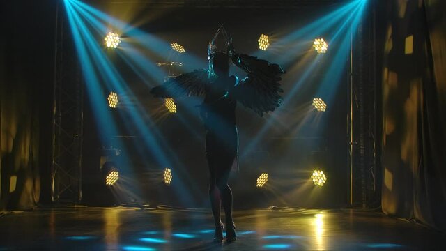 A young actress in the image of the Greek goddess Artemis poses on stage in the rays of magical light. Silhouette of a beautiful woman with golden wings and a headdress with feathers. Slow motion.