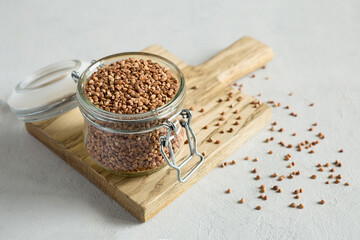Buckwheat uncooked grains in glass jar on the wooden cutting board close up on the gray background. Healthy, dietary food. Organic, carbohydrate product. Food storage