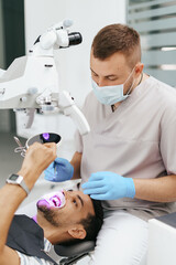 Naklejka na ściany i meble Young man with patient bib on a dental chair and a dentist who sits next to him. He looks on his teeth using a dental microscope and holds a dental bur and a mirror.