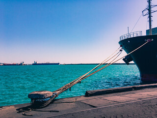 A mooring bollard with a fixed cable and the bow of a ship against the backdrop of a seascape with ships on the horizon under a blue cloudless sky.
