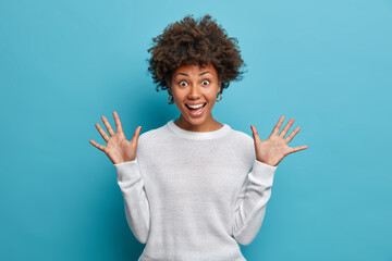 Amused pretty girl with curly Afro hair, raises palms, has cheerful expression, smiles broadly,...
