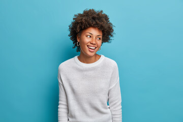 Joyful dark skinned young woman with broad smiles, perfect teeth, feels carefree and enthusiastic, looks happily aside, stands entertained, wears casual white jumper, isolated on blue background