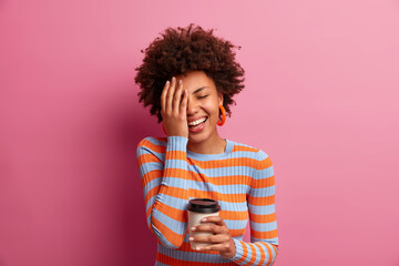 Indoor shot of good looking curly haired woman makes face plam, closes eyes, laughs positively, smiles broadly, has happy talk with friend, enjoys caffeine beverage in morning, dressed casually