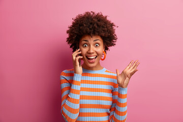 Emotional cheerful girl with curly hair calls best friend, raises hand and has joyful expression, wears striped casual turtleneck, poses against pink background, hears excellent news, being very glad