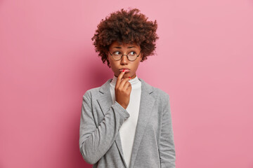 Fototapeta na wymiar Sad discontent curly woman executive worker looks unhappily aside, purses lips, expresses regret, has problem, needs good advice, dressed formally, isolated on pink wall, thinks of concequences