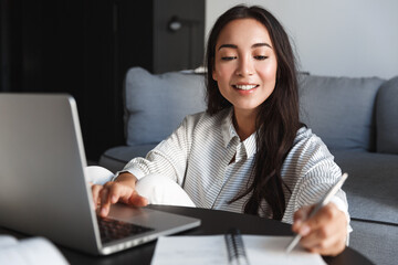 Image of beautiful asian girl studying remote from home, writing down lecture while using virtual...