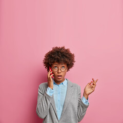 Surprised beautiful Afro American woman employee talks via mobile phone and looks above, raises hand, wears round glasses, formal clothes, isolated on pink background, copy space for your text