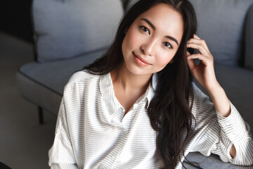 Close-up of attractive brunette asian girl sitting on floor near sofa, looking dreamy and sensual at camera with tender smile. Woman resting at home in front of TV, relaxing and watching movies