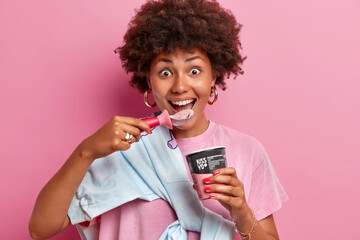 Close up shot of cheerful funny teenage girl with curly hairstyle enjoys eating cold frozen dessert, eats delicious ice cream of strawberry flavor from spoon, stands indoor, pink background.