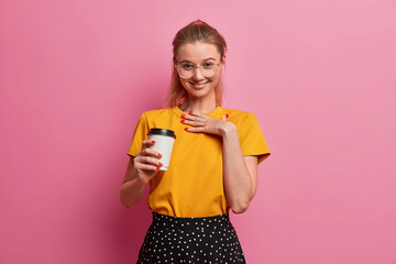 Horizontal shot of pretty young European girl giggles positively, cheers over something, drinks takeout coffee, smiles from satisfaction, has fun in merry company, communicates with friends.