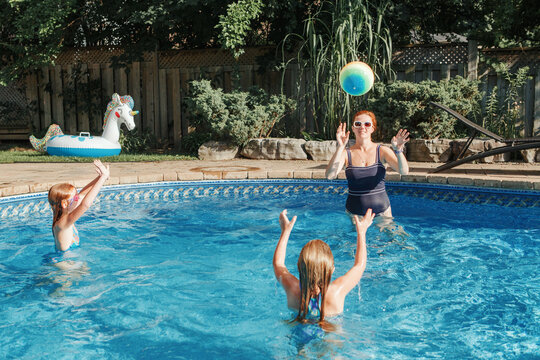Mother playing ball with daughters children in swimming pool on home backyard. Mom and sisters siblings having fun in swimming pool together. Summer outdoors water activity for family and kids.