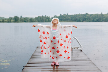 Girl child in rain poncho with red maple leaves standing on wooden lake dock. Kid rasing arms up...